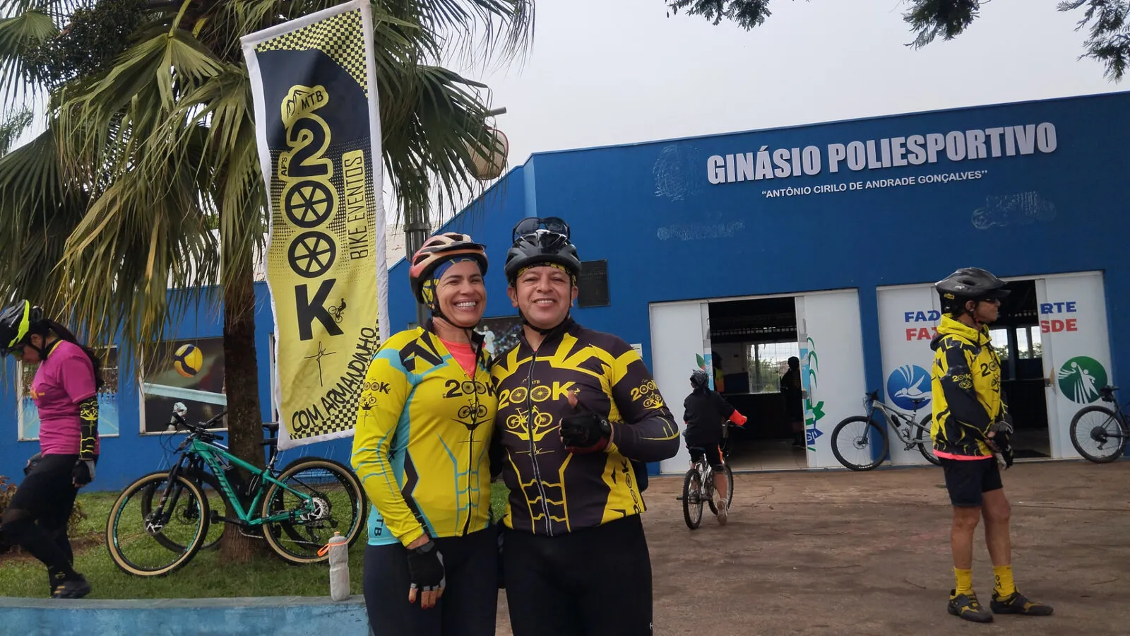 Dois ciclistas com camisas amarelas do 200K sorrindo em frente a um ginásio poliesportivo, ao lado de uma bandeira do grupo
