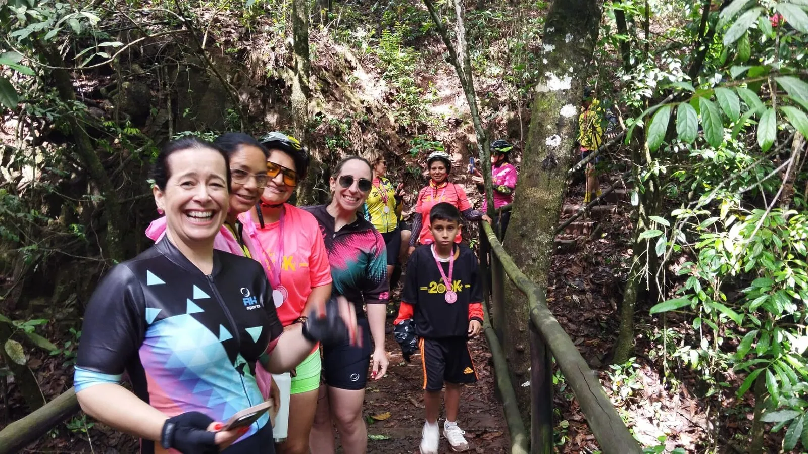 Ciclistas sorrindo em uma trilha no meio da mata, com uma criança de medalha no pescoço