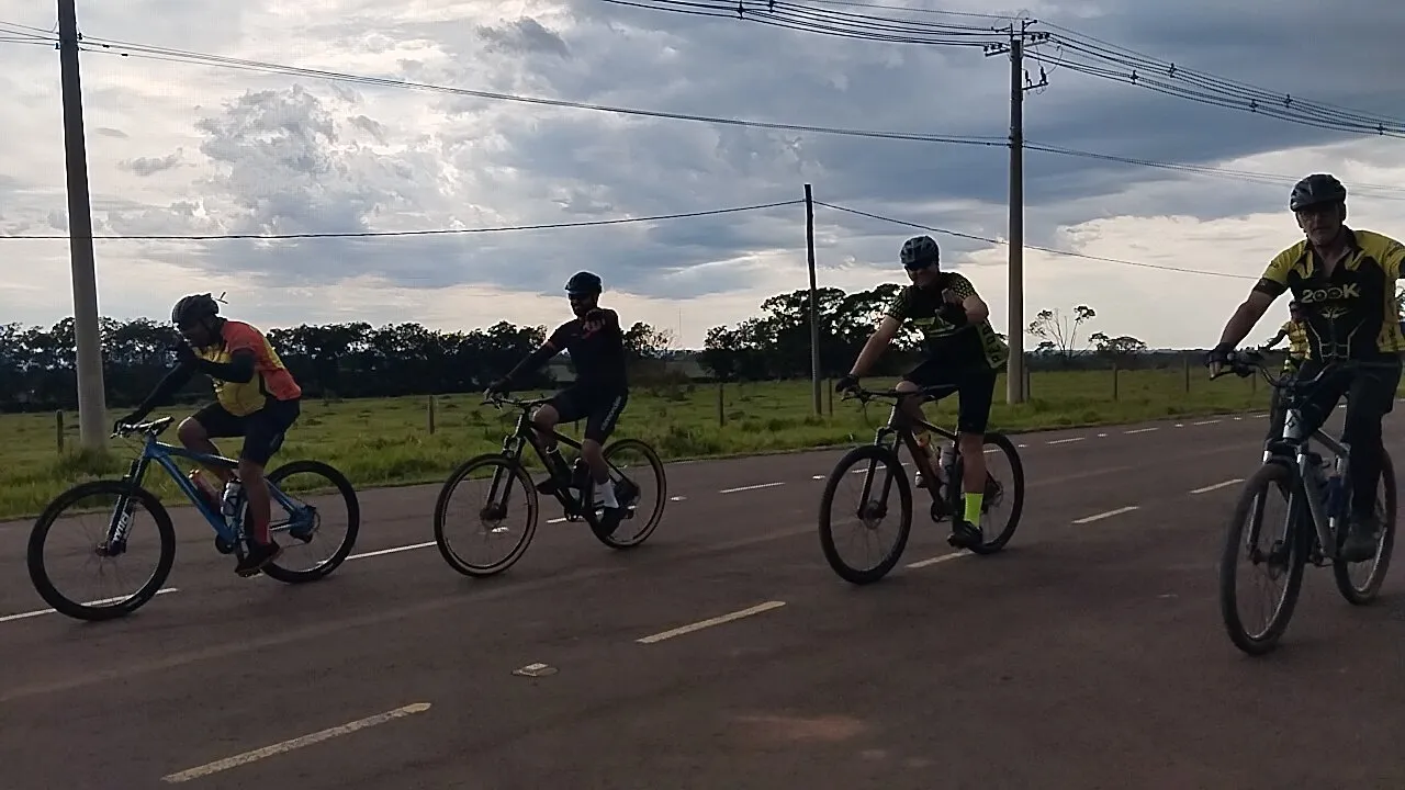 Quatro ciclistas pedalando lado a lado em uma estrada de asfalto com céu nublado