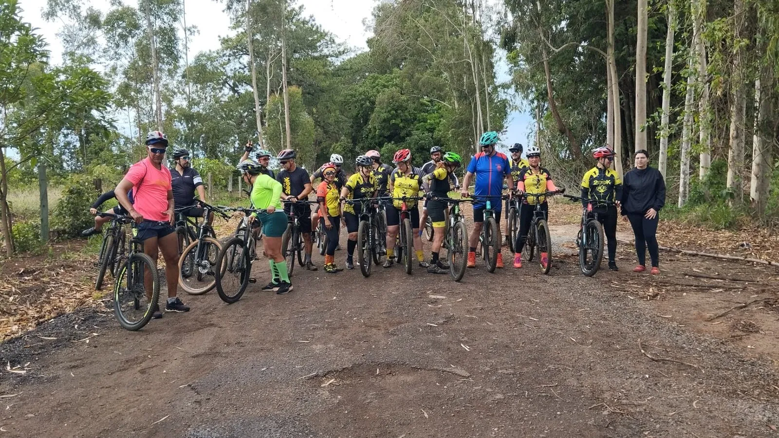 Grupo grande de ciclistas parados com as bikes em uma estrada de terra entre eucaliptos