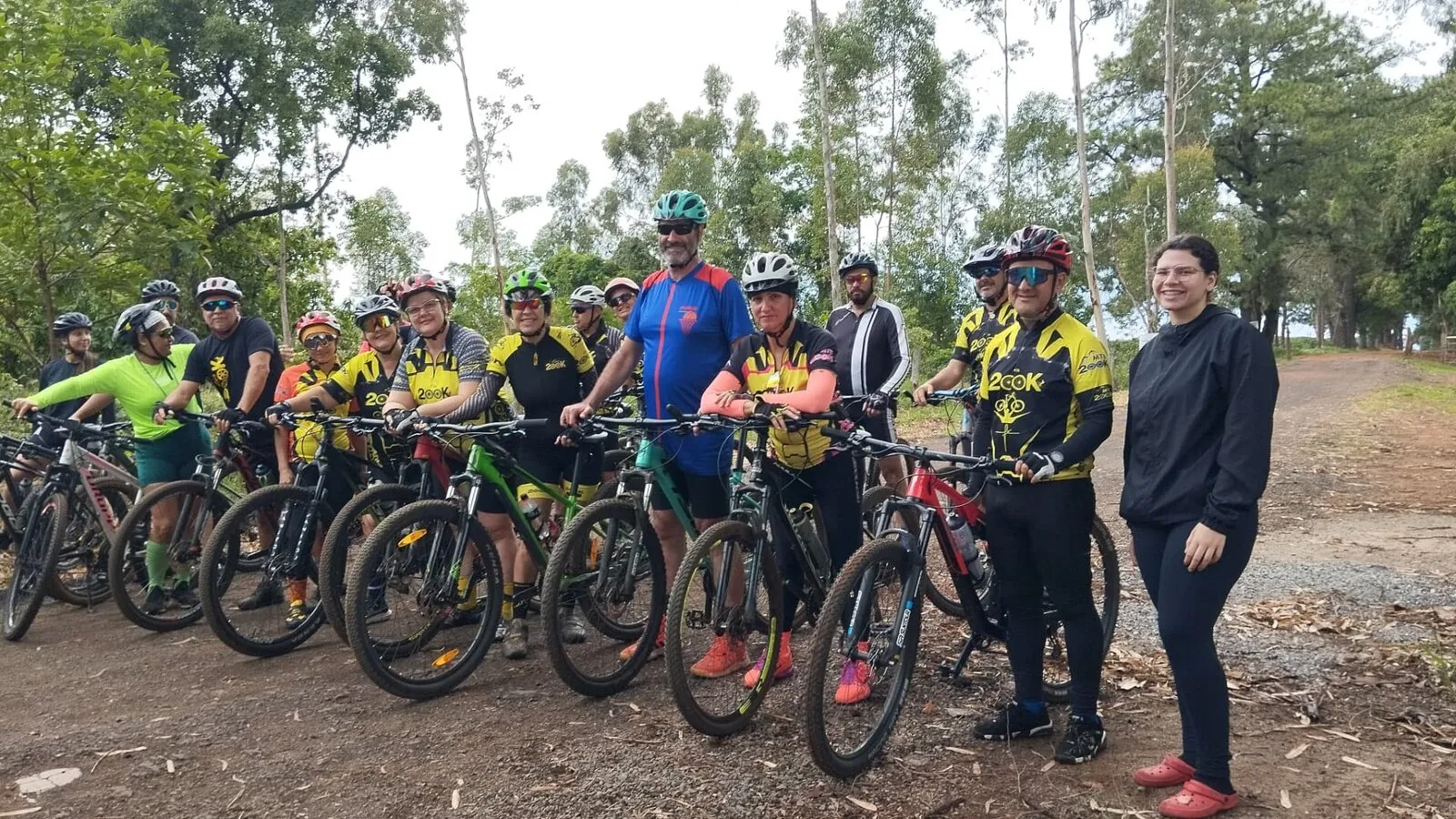 Ciclistas do grupo enfileirados com as bikes em uma estrada de terra, com árvores ao fundo